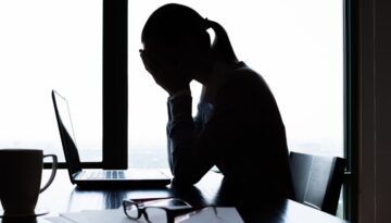 Woman sitting by a laptop with her hands over her face looking stressed.