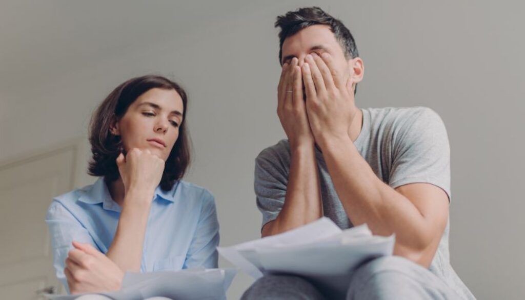 Man and Woman have paperwork on their laps. The man covers his face with his hands and the woman looks thoughtfully over at the man's papers.