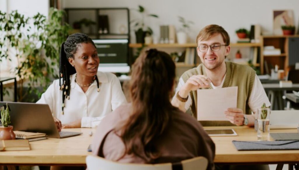 A man and woman sit across from another woman in a hiring interview. The man holds a piece of paper and is smiling.