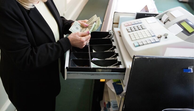 An employee counting twenty dollar bills in a cash register.