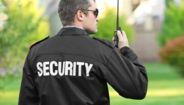 Security guard stands outside and is speaking into a handheld walkie-talkie.