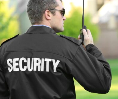 Security guard stands outside and is speaking into a handheld walkie-talkie.