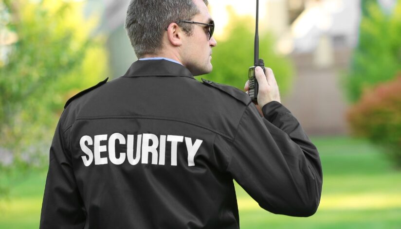 Security guard stands outside and is speaking into a handheld walkie-talkie.