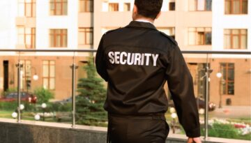 Security Guard faces away from lens, looking over a glass balcony towards green plants.