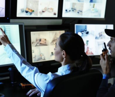 Two people look at screens with surveillance footage. The woman on the left points at a screen while the man on the right speaks into a walkie-talkie.